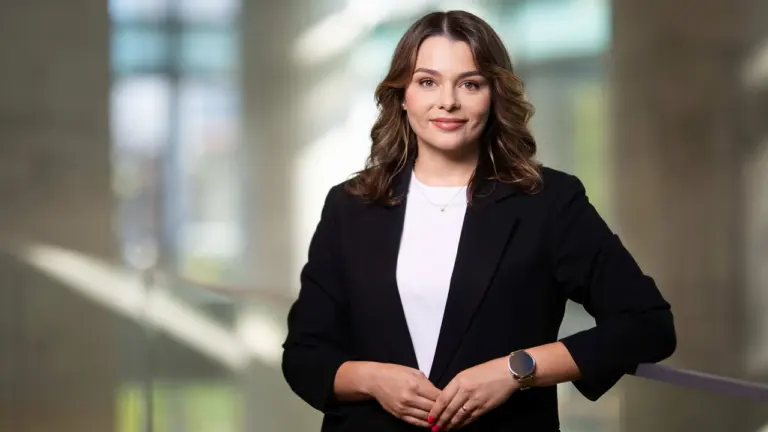 Jauna moteris šviesiais banguotais plaukais vilki juodą švarką ir stovi šviesiame biuro fone.