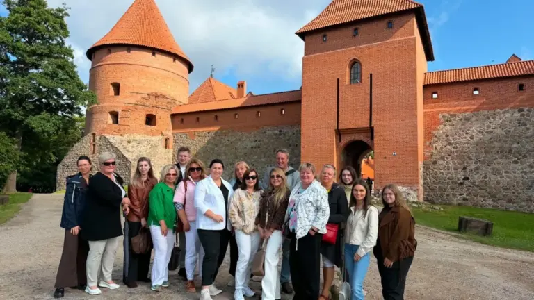 Grupė besišypsančių žmonių stovi prie Trakų pilies raudonų plytų sienų ir bokštų fone.