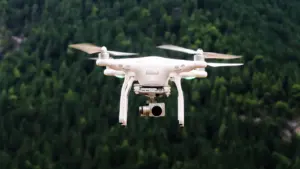 Baltas dronas skrenda virš tankaus miško Lietuvos kraštovaizdyje