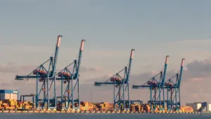 Dideli pramoniniai kranai Klaipėdos uosto terminale, simbolizuojantys uosto infrastruktūros plėtrą ir modernizavimą.