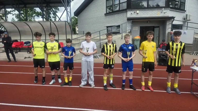 Jaunieji futbolininkai su sportine apranga stovi stadione ir rankose laiko laimėtus apdovanojimus bei taures.